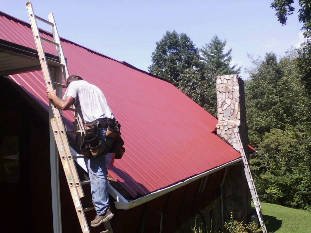 Roofer on a ladder beside a red metal roof and stone chimney — Soffit & Fascia Repair by Roofing Brothers in Newkirk